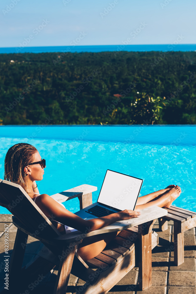 Young woman in sunglasses resting on sunbed near blue swimming pool and working freelance at modern latop computer with blank copy space screen for advertising information content enjoying summertime
