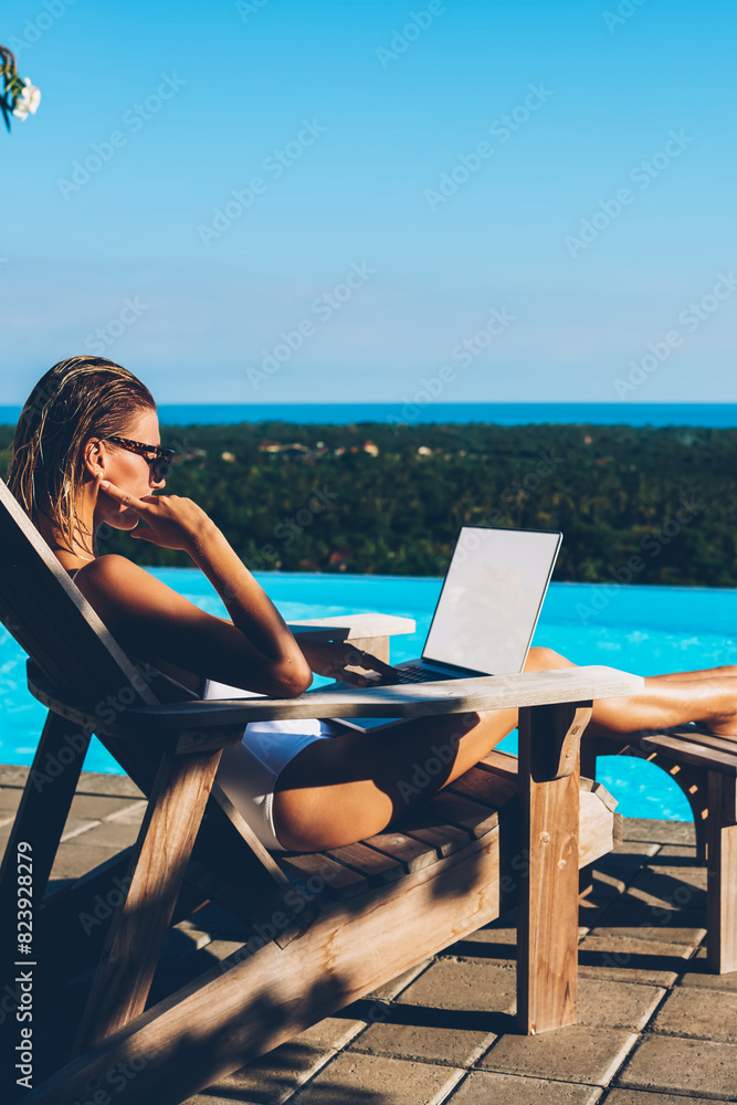 Pensive slim young woman working freelance at netbook with blank screen area while resting on sunbed near swimming blue poor enjoying summer resort.Tourist reading news on laptop while sunbathing