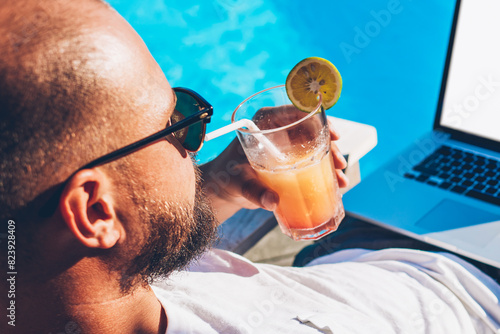 Young man checking email on smartphone and synchronizing media from phone on laptop with blank copy cpace screen for your advertising information content while drinking cocktail lying on sunbed photo