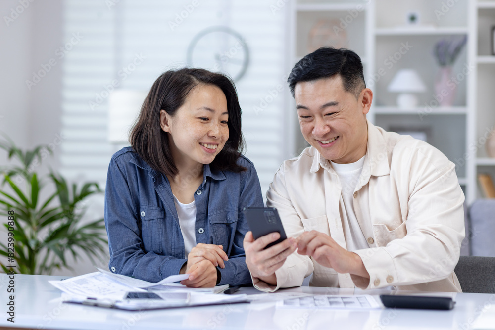 Φωτογραφία Smiling couple managing their finances together at home using a smartphone, looking at bills and papers, in a bright and modern living room