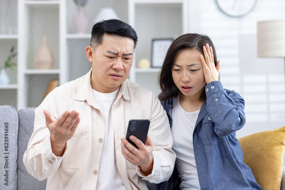 Concerned couple reacting to something on a smartphone screen, showing surprised and confused expressions, sitting in a living room Concerned couple reacting to something on a smartphone screen, showing surprised and confused expressions, sitting in a living room
