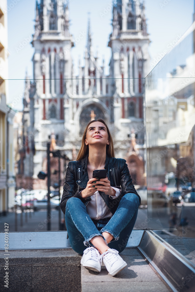 Fototapeta premium Beautiful young girl in casual clothes is holding a smartphone, looking upward and smiling while resting in city center
