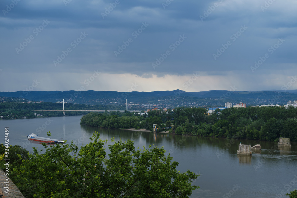 Serbia - Beautiful Panoramic view of Novi Sad and bridge across Danube River. View from Petrovaradin fortress. A large barge ship is sailing on the river. It's raining in mountains in the distance.