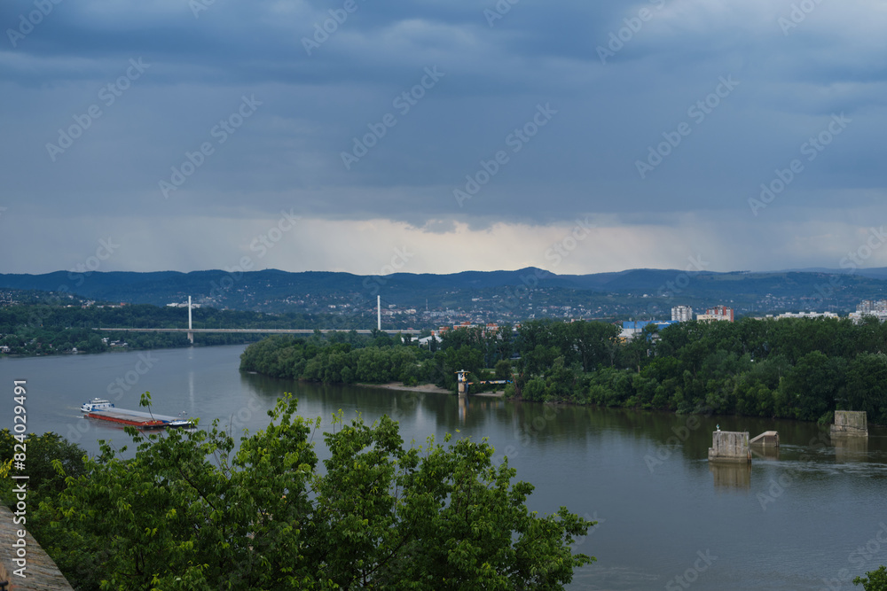 Serbia - Beautiful Panoramic view of Novi Sad and bridge across Danube River. View from Petrovaradin fortress. A large barge ship is sailing on the river. It's raining in mountains in the distance. Serbia - Beautiful Panoramic view of Novi Sad and bridge across Danube River. View from Petrovaradin fortress. A large barge ship is sailing on the river. It's raining in mountains in the distance.
