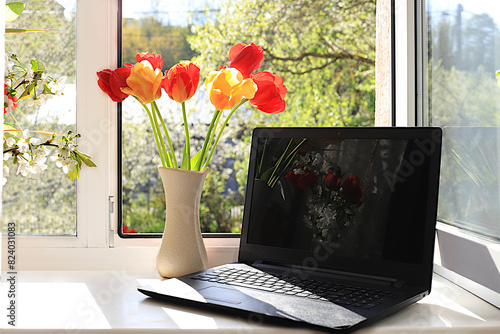Computer and tulip flowers in a vase on a sunny window. The concept of home coziness, comfort and home office. Computer mobility and healthy lifestyle, selective focus..