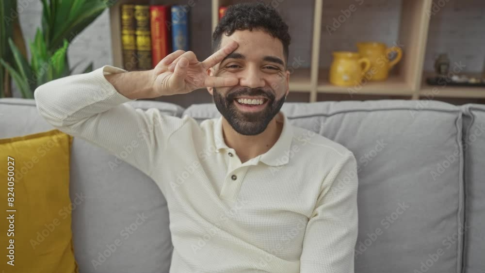 Young hispanic man, cheerful and smiling, showing peace victory symbol with fingers on face at home Young hispanic man, cheerful and smiling, showing peace victory symbol with fingers on face at home