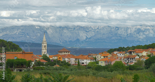 Windy day in coastal island town nestled against dramatic mountain range under cloudy sky and rippled blue Adriatic Sea. Tourist village by the rough and wavy sea after passage of the summer front.