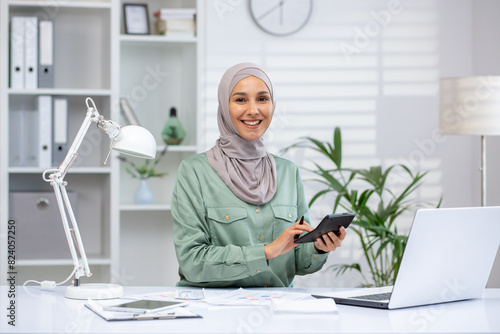 Portrait of successful and happy businesswoman financier, woman in hijab smiling and looking at camera, using calculator at work, accountant working with papers using laptop