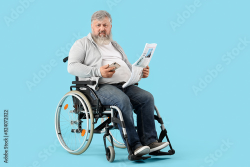 Portrait of sad senior man in wheelchair with mobile phone reading newspaper on blue background
