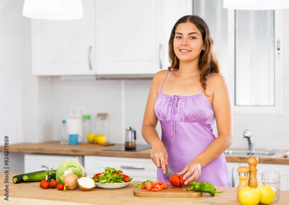 Smiling young woman in nightie cooking vegetable salad in kitchen at home.