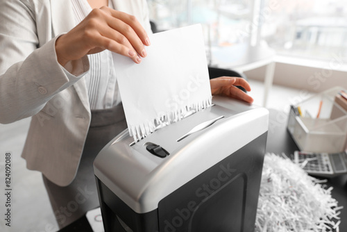 Young secretary destroying paper sheet using shredder in office, closeup