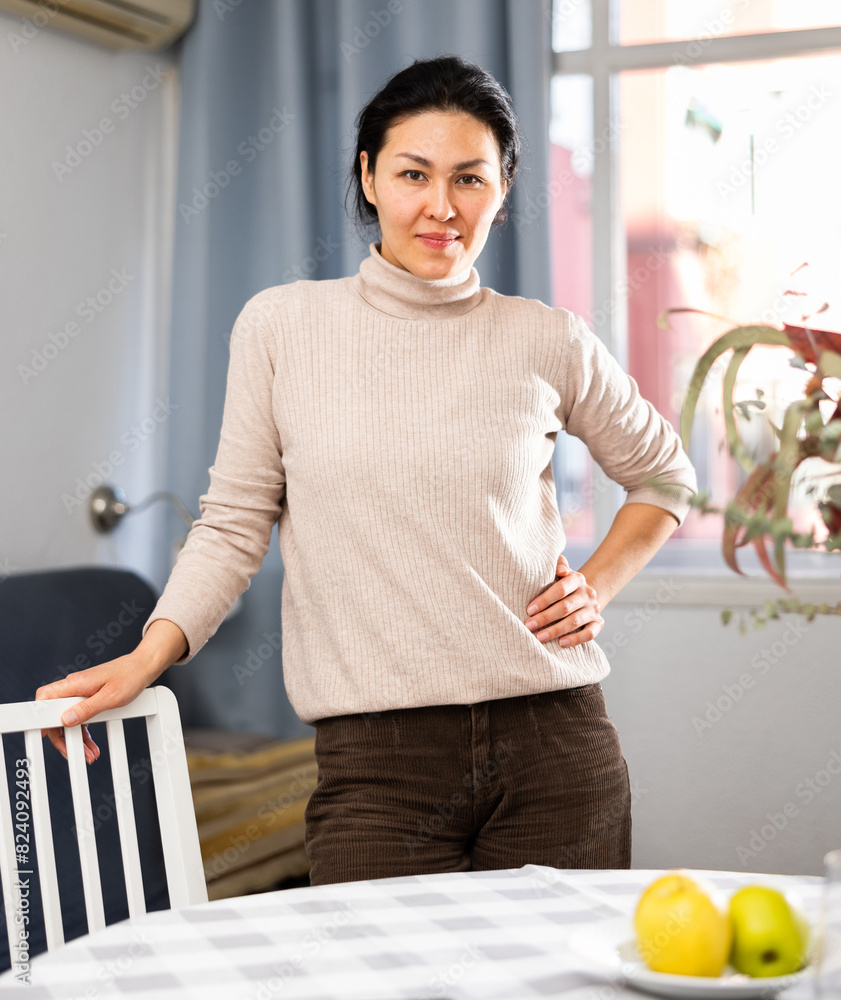 Obraz premium Portrait of a positive asian woman standing near a table in a room and posing for the camera