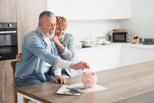 Mature couple putting coin into piggy bank in kitchen