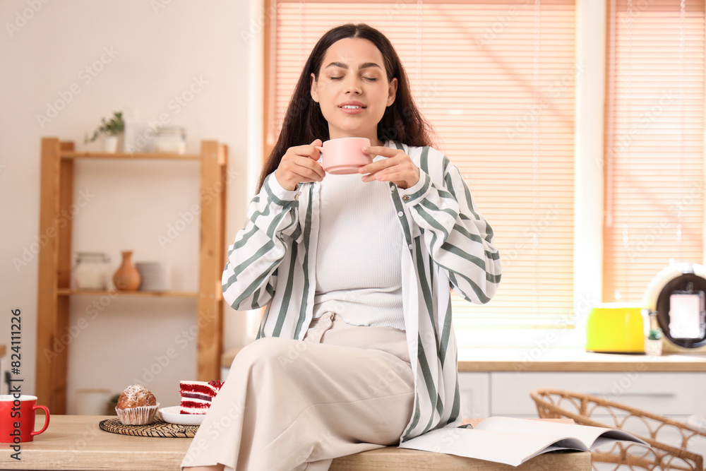 Young happy woman with cup of fresh coffee and desserts reading magazine in kitchen Young happy woman with cup of fresh coffee and desserts reading magazine in kitchen