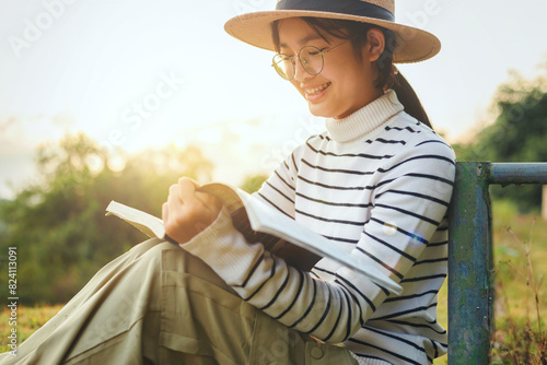 Cute girl with eyeglasses and hat sitting on grass and reading book in park sunset.Side view .
