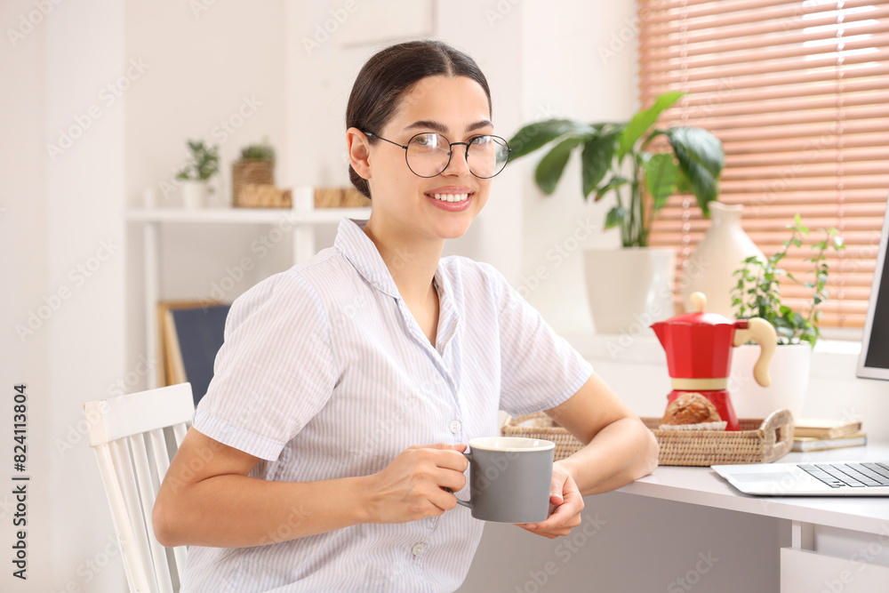 Pretty young woman with cup of hot coffee and laptop sitting at table in light room Pretty young woman with cup of hot coffee and laptop sitting at table in light room