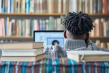 A student with short dreadlocks, wearing a grey sweater, is seen from behind while studying in a library.