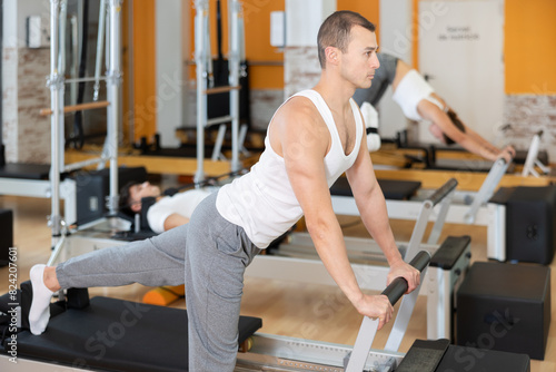 Man athlete works out in gym and performs set of exercises to stretch leg muscles with help of reformer. Male visitor to fitness pilates center is doing exercises in background. © JackF