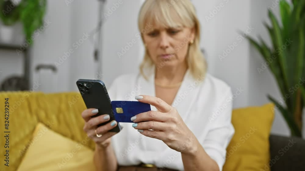 A blonde woman examines her credit card while using a smartphone in a cozy living room setting. A blonde woman examines her credit card while using a smartphone in a cozy living room setting.