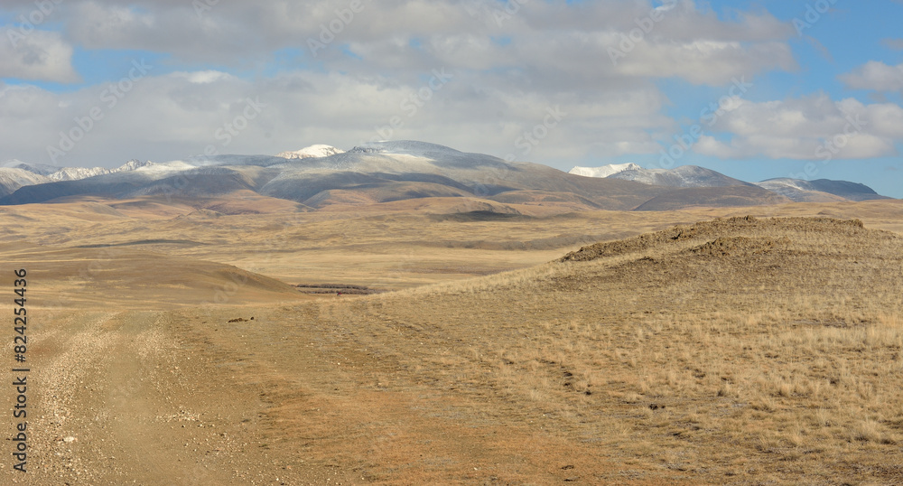 A field road going through the hilly autumn steppe towards high and snow-covered mountain ranges.