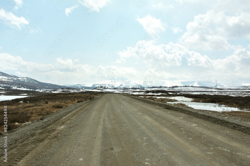 A muddy gravel road crosses a huge snow covered clearing surrounded by high mountain ranges under a cloudy spring sky. A muddy gravel road crosses a huge snow covered clearing surrounded by high mountain ranges under a cloudy spring sky.