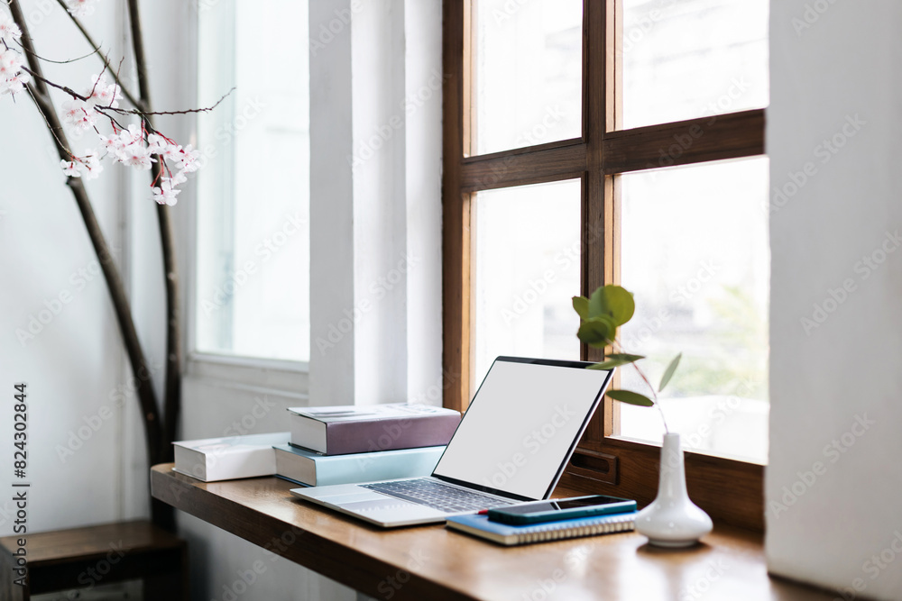 Fototapeta premium Comfortable workplace on wooden counter by the window and laptop, smart phone, note book and pen. vase with small tree inside and a book for read
