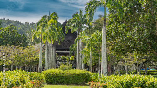 Landscape design of a tropical park. Tall royal palm trees Roystonea regia grow in two rows. Lush green bushes, deciduous trees all around. A building with a triangular roof is visible in the distance photo