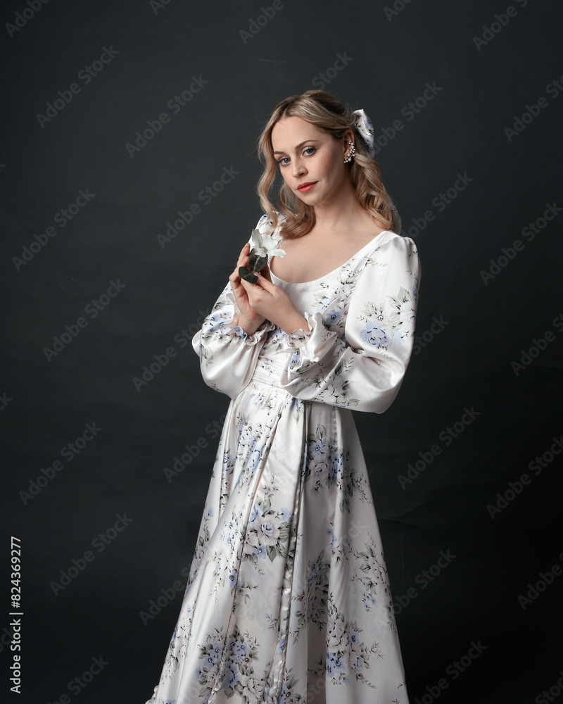 Obraz premium close up portrait of beautiful blonde female model wearing romantic historical gown of white silk with floral pattern and bow. Elegant standing pose, isolated on dark studio background.