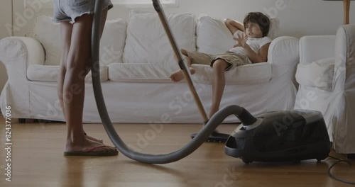 A mother vacuums the living room while her young son watches from the couch, appearing bored and uninterested. photo
