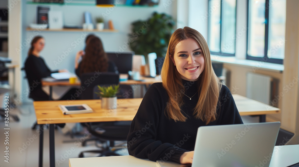 A smiling woman sitting at a desk in a modern, open-plan office working on a laptop A smiling woman sitting at a desk in a modern, open-plan office working on a laptop