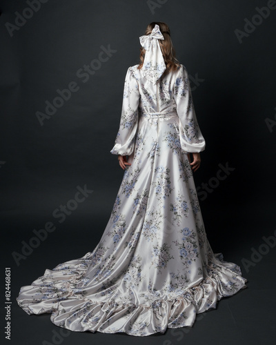 portrait of beautiful blonde female model wearing romantic historical gown white bridal floral pattern. standing pose running away from camera with flowing silk skirt. isolated dark studio background.