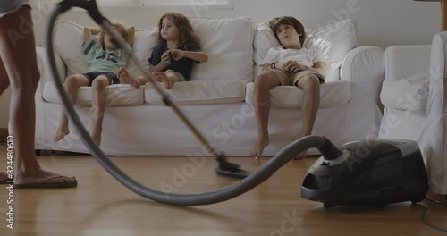 A mother vacuums the living room while her young son watches from the couch, appearing bored and uninterested.