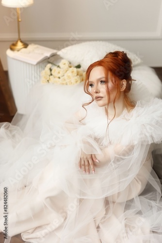 Graceful young woman with fiery red hair and flawless makeup in a stunning white bridal gown. She poses elegantly in a photo studio with a classic light interior, exuding a serene charm