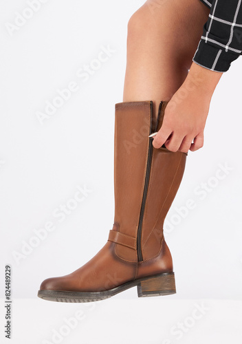 Young woman wearing a stylish brown leather knee-high boot, zipping up the side closure, fine stitching, and a sleek, smooth finish, clean white background, quality craftsmanship and elegant design.
