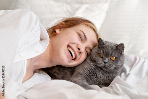 Happy teenager girl laughing laying on bed together with gray cat, people and pets affection and care