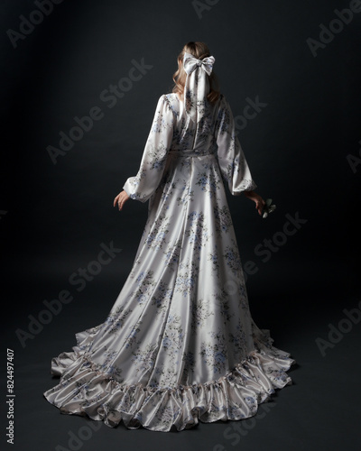 portrait of beautiful blonde female model wearing romantic historical gown white bridal floral pattern. standing pose running away from camera with flowing silk skirt. isolated dark studio background.