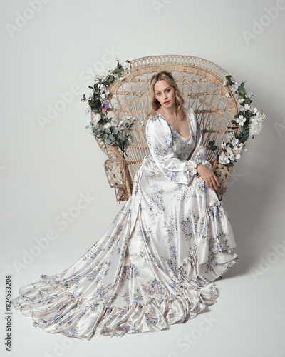 portrait of beautiful blonde female model wearing romantic historical white bridal gown. sitting pose on floral Peacock Throne chair with flowing silk skirt. isolated dark studio background.