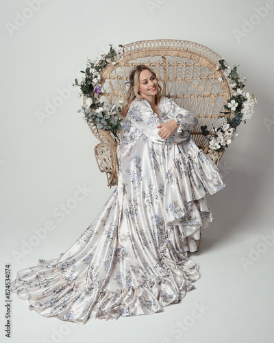 portrait of beautiful blonde female model wearing romantic historical white bridal gown. sitting pose on floral Peacock Throne chair with flowing silk skirt. isolated dark studio background.