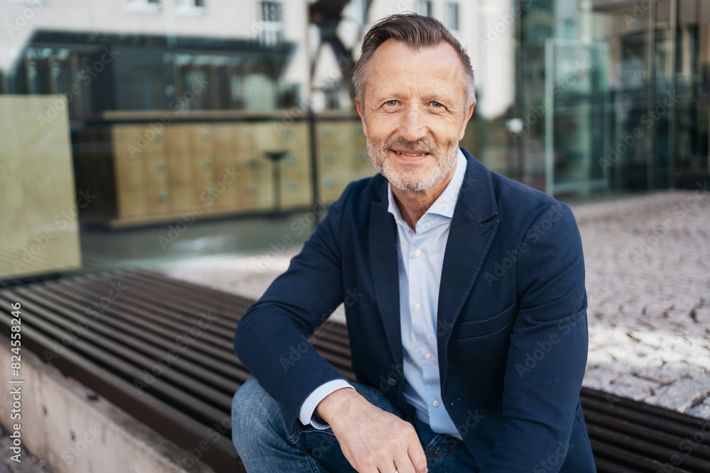 A confident mature man smiling while seated outdoors in business casual attire. Urban background suggests a professional environment. A confident mature man smiling while seated outdoors in business casual attire. Urban background suggests a professional environment.