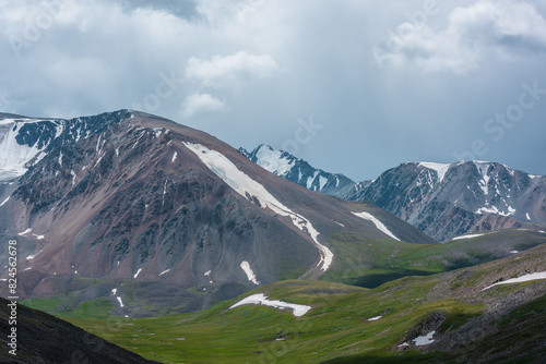 Dramatic overcast view to beautiful green alpine valley among stony and grassy hills, big snowy range and part of mountain with snow-capped sharp pinnacle far away in center under rain gray cloudy sky