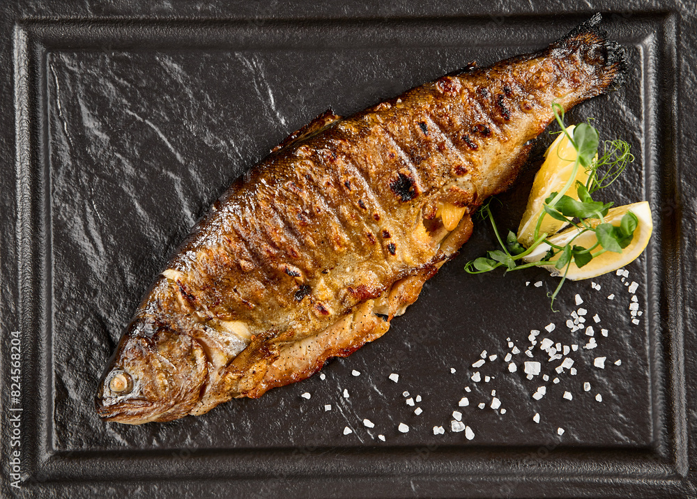 Grilled rainbow trout with a lemon wedge and fresh herbs on a dark stone plate Grilled rainbow trout with a lemon wedge and fresh herbs on a dark stone plate