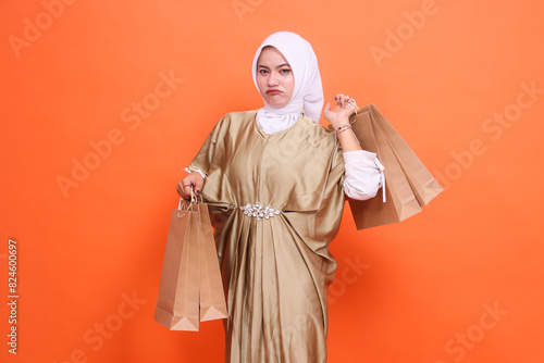 young indonesia woman sullenly carries and lifts a paper bag containing groceries wearing a kaftan Muslim dress with a hijab, isolated on an orange background. Fashion concept, transaction, promotion photo