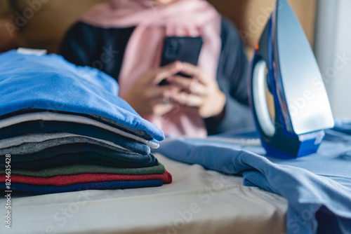A stack of clothes that has been ironed, with a background of women with a headscarf playing with a phone, concept of tacking break while doing a chores