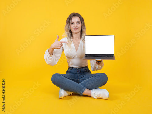 Pointing laptop screen, full body size front view young caucasian woman sit floor holding pointing laptop screen. Empty blank white display, mock up. Yellow background. Copy space. Advertisement idea.