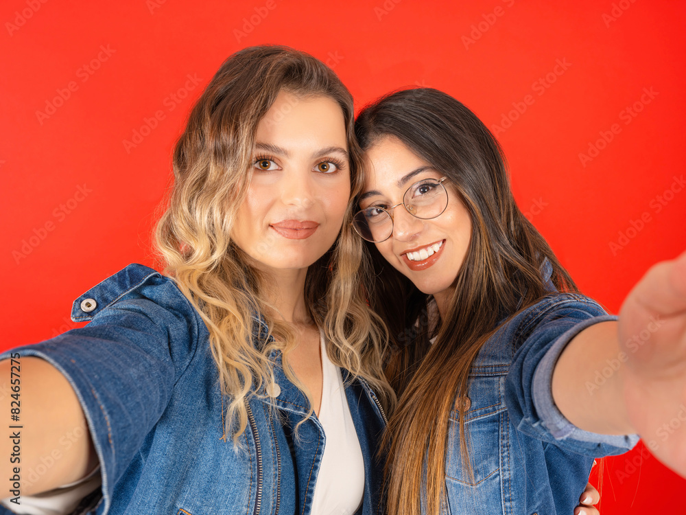 Making selfie, cute beautiful two young pretty best friends girls having fun making selfie. Positive smiling emotions. Standing over red studio background. Wear denim jackets, people lifestyle concept Making selfie, cute beautiful two young pretty best friends girls having fun making selfie. Positive smiling emotions. Standing over red studio background. Wear denim jackets, people lifestyle concept