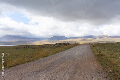 A gravel road through autumn fields leading to a ridge of mountains covered with dense clouds.