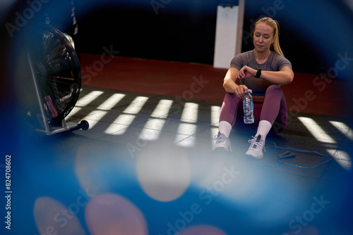 Sporty woman sitting on the gym floor, checking her smartwatch after a workout, with a water bottle and jump rope beside her.