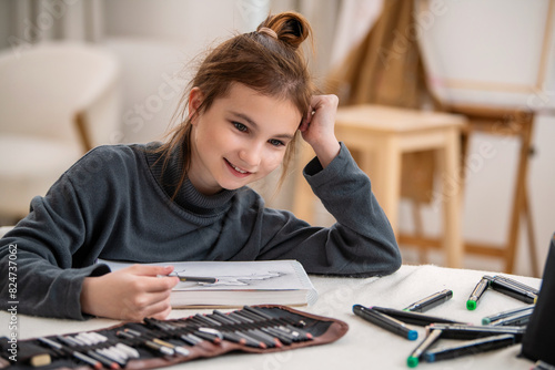 A young girl is sitting at a table with a notebook and a bag of pencils.