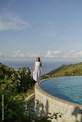 Romantic girl in long white dress enjoying rest at exotic resort. with nature and ocean view Outdoor portrait of female model posing near  infinity pool during vacation on Bali island