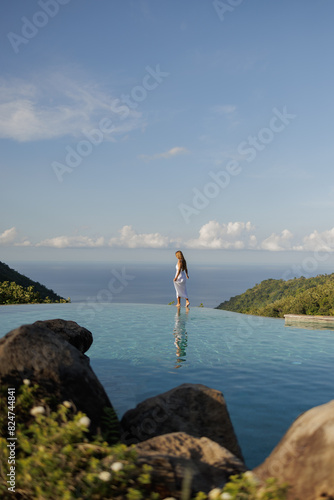 Asian woman in white dress walk on the edge of infinity pool above the sea with nature view. Young woman traveler relaxing at sky pool and looking at the ocean